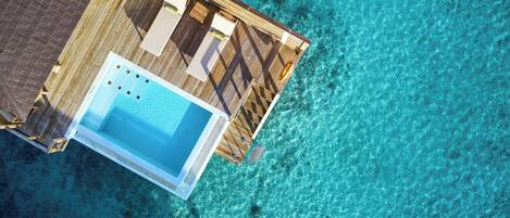 An aerial view of a wooden deck with a pool and lounge area, surrounded by clear turquoise water.