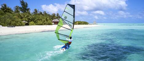 A person windsurfing on turquoise waters with a clear sky and palm trees in the background.