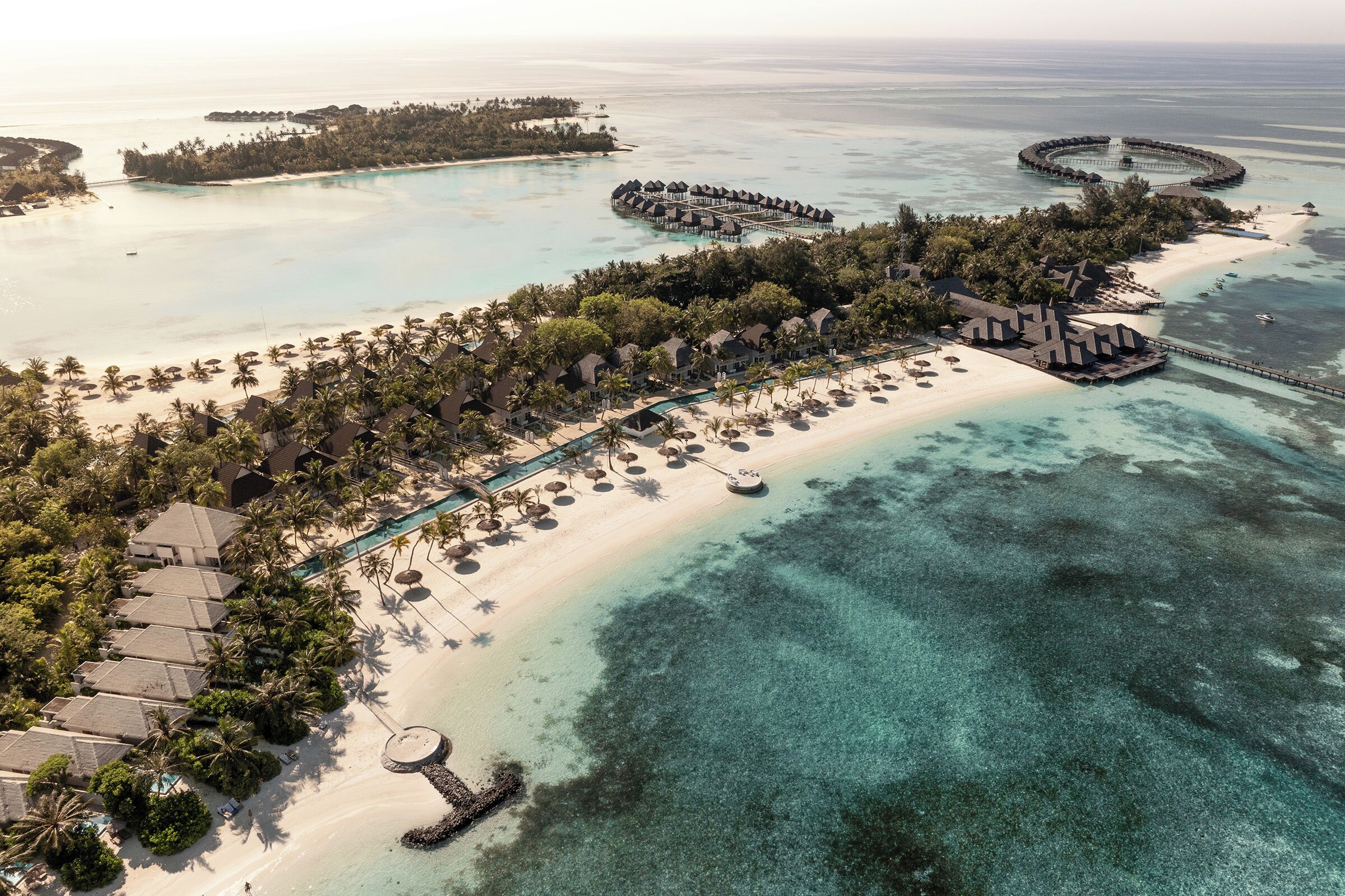 Vue aérienne d’un complexe hôtelier doté d’une plage, de bungalows sur pilotis et d’une structure circulaire au milieu de l’eau.
