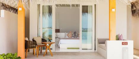 A hotel room with a balcony, a bed with pillows, a table with chairs, and a thatched roof.