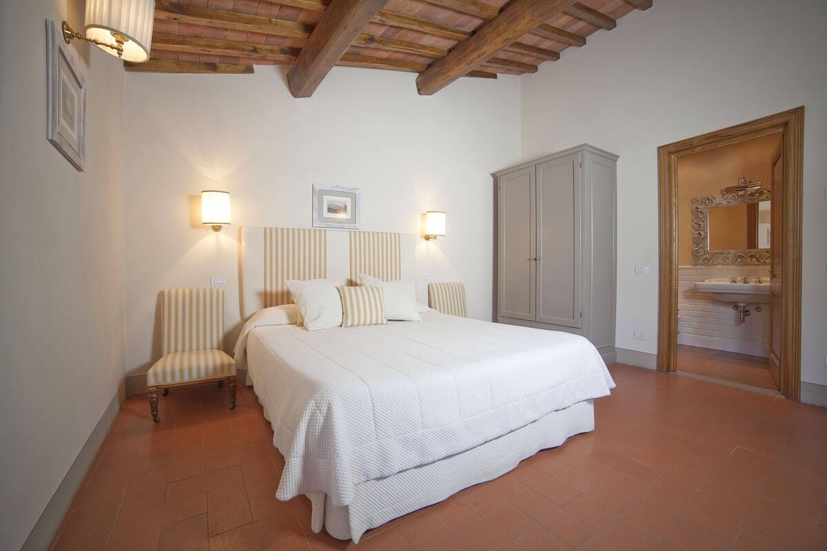 A bedroom with a large bed, two bedside lamps, a chair, a wardrobe, and a bathroom with a mirror and sink.