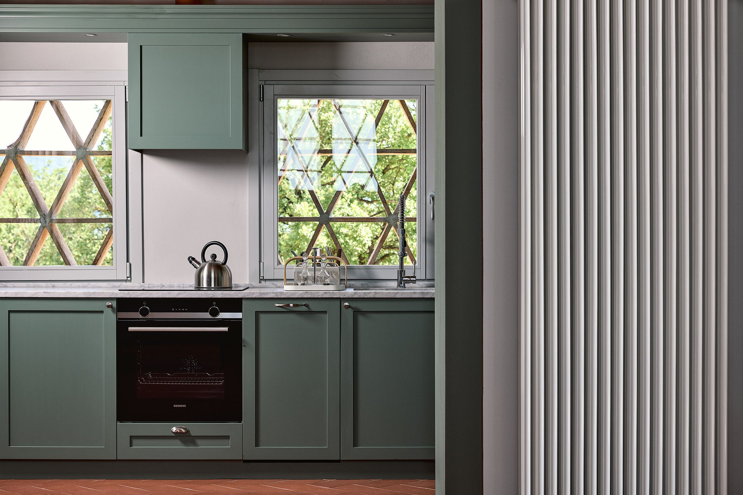 A kitchen with a built-in oven, a kettle, and a window with a view of greenery.