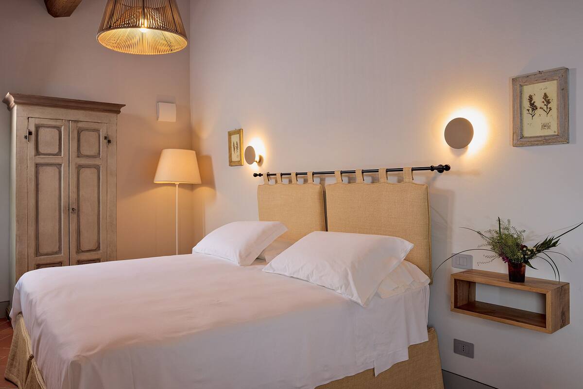 A neatly made bed with white linens, a bedside lamp, and a small wooden shelf with a plant.
