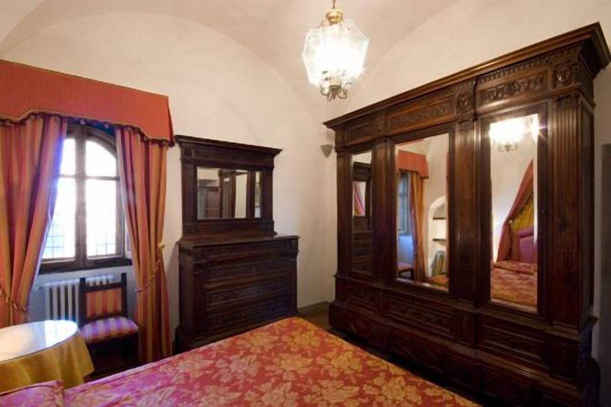 A bedroom with a large wooden wardrobe, a bed with a floral bedspread, a window with curtains, and a chandelier.