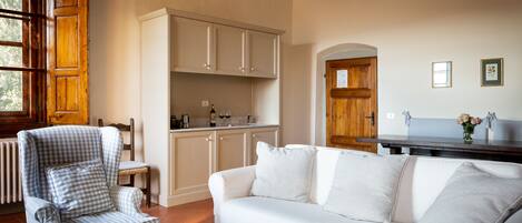 A living room with a white sofa, a checkered armchair, a wooden coffee table, and a kitchen area with white cabinets and a wooden door.