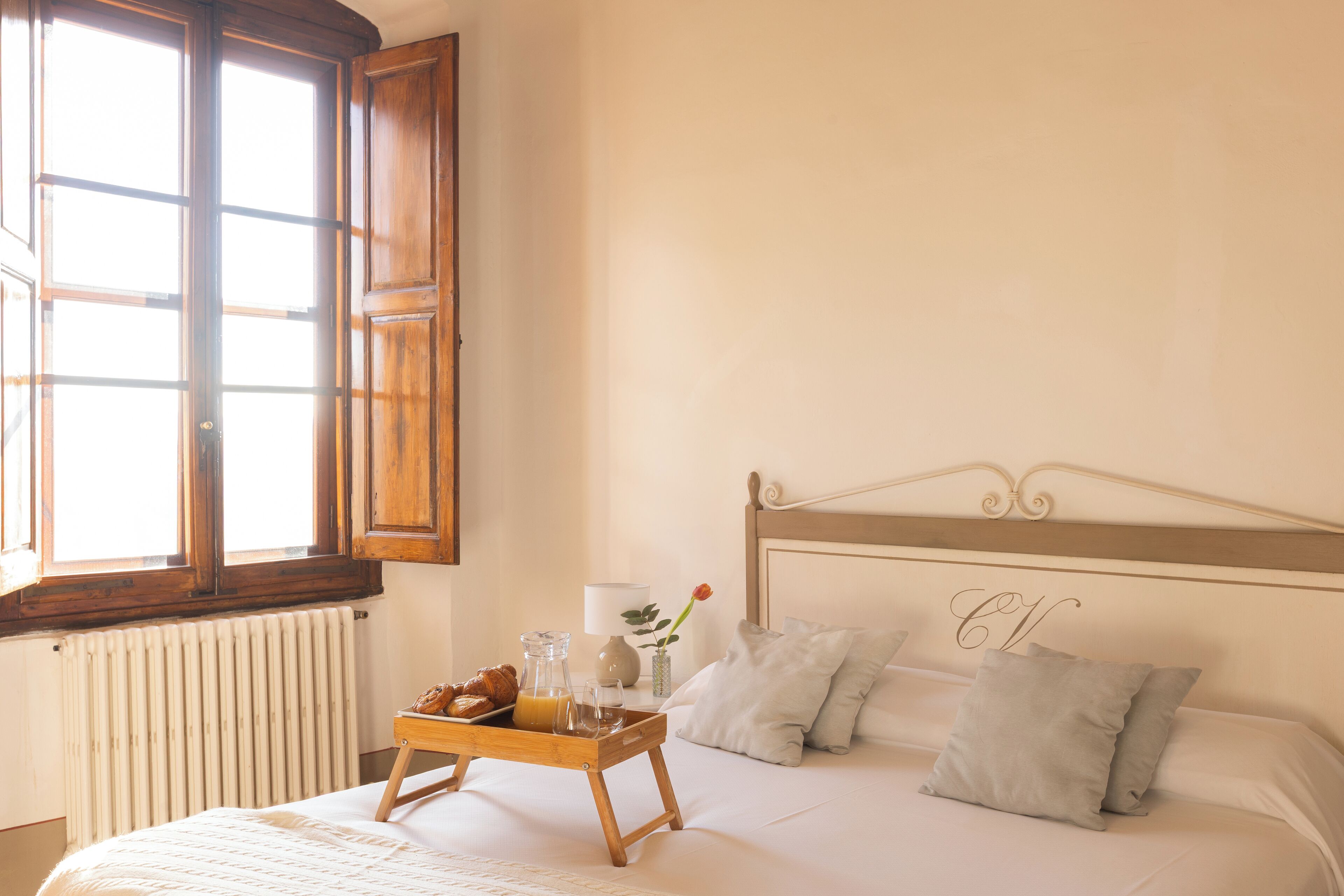 Ein ordentlich bezogenes Bett mit einem Frühstücksbrettchen, einer Vase mit einer Blume und einem Fenster mit hölzernen Rollläden.