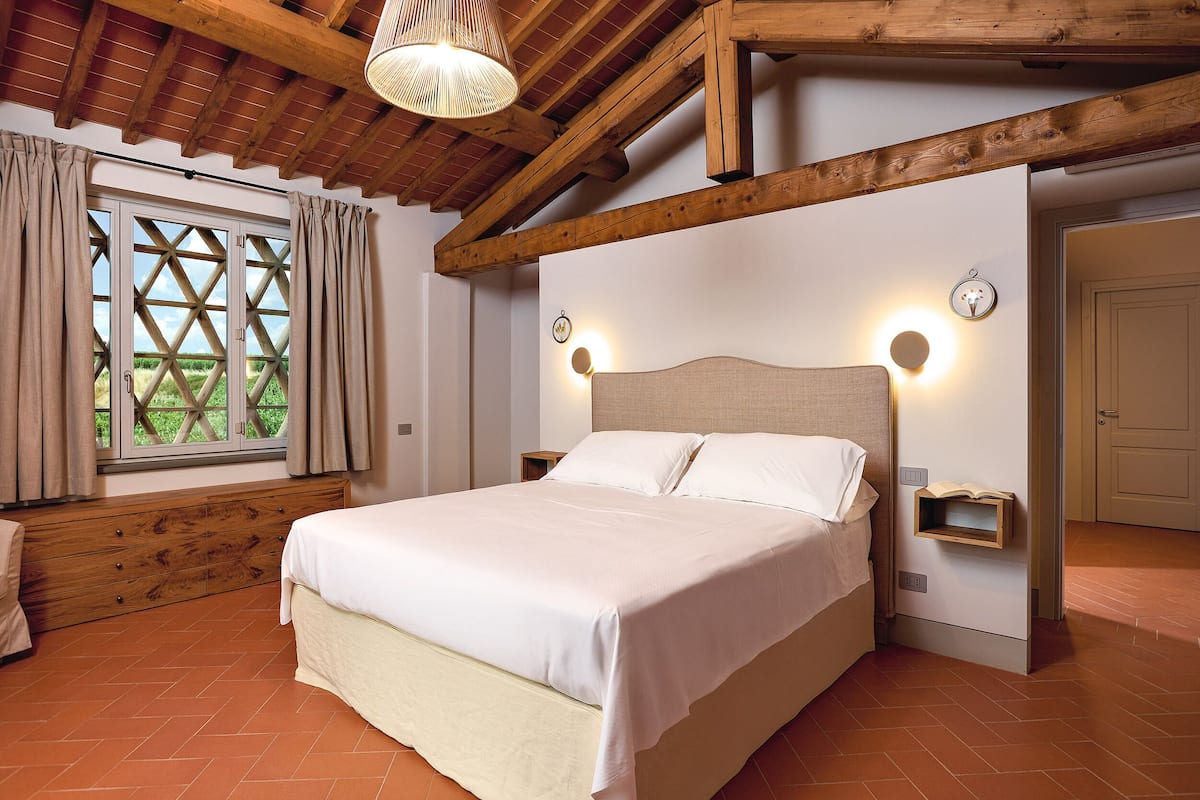 A bedroom with a bed, wooden beams, a window with curtains, and wall-mounted lights.