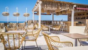 A beachside outdoor seating area with wicker chairs and tables, a bar with bottles, and a view of the sea. - Villa Elite by Checkin (Hersonissos)