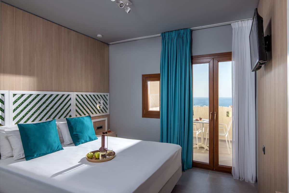 Habitación de hotel con cama, ventana con cortinas, vista al mar y balcón con mobiliario exterior.