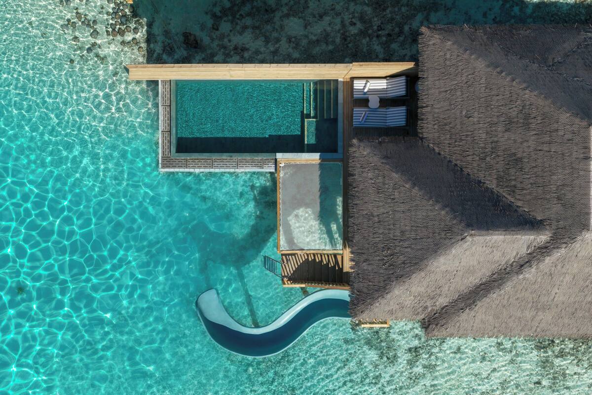 Eine Vogelperspektive zeigt einen luxuriösen Urlaubsort mit einem türkisfarbenen Pool, einer Holzterrasse und einem Sandstrand.