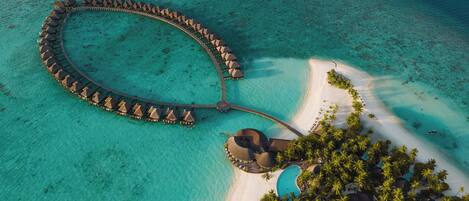 An aerial view of a resort with overwater bungalows arranged in a semi-circle, connected by a bridge, and a separate area with a pool and palm trees.