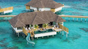 Dos bungalows sobre el agua con techos de paja, una piscina y un tobogán, unidos por un paseo de madera sobre aguas turquesas cristalinas.