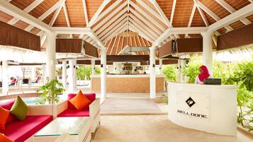 A beachside restaurant with a thatched roof, outdoor seating, and a reception desk.