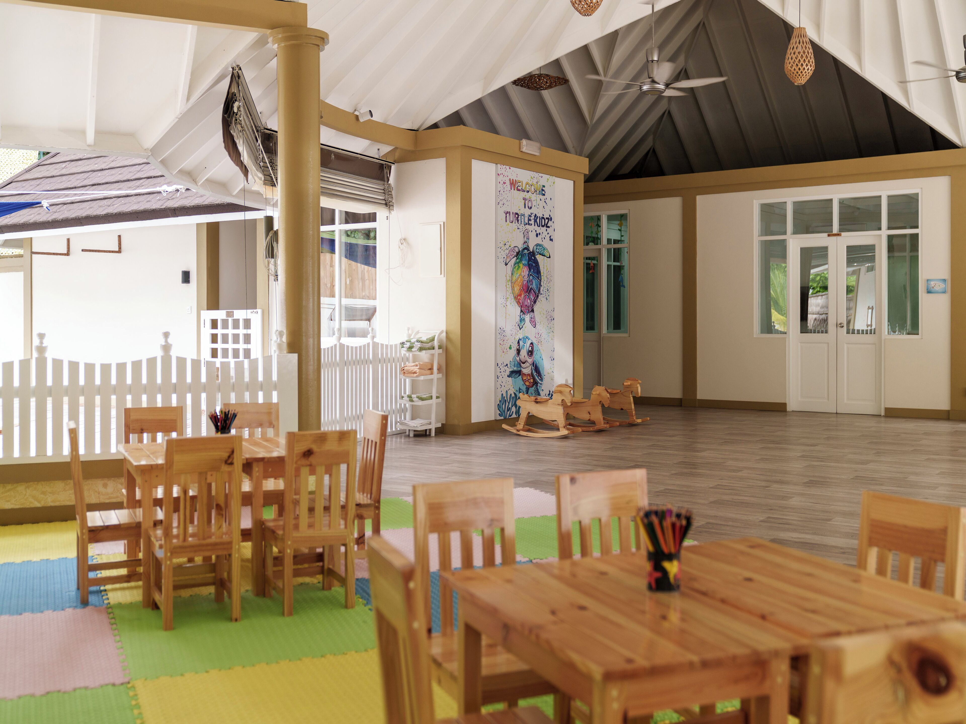 A spacious indoor area with wooden tables and chairs, a white picket fence, and a mural on the wall.