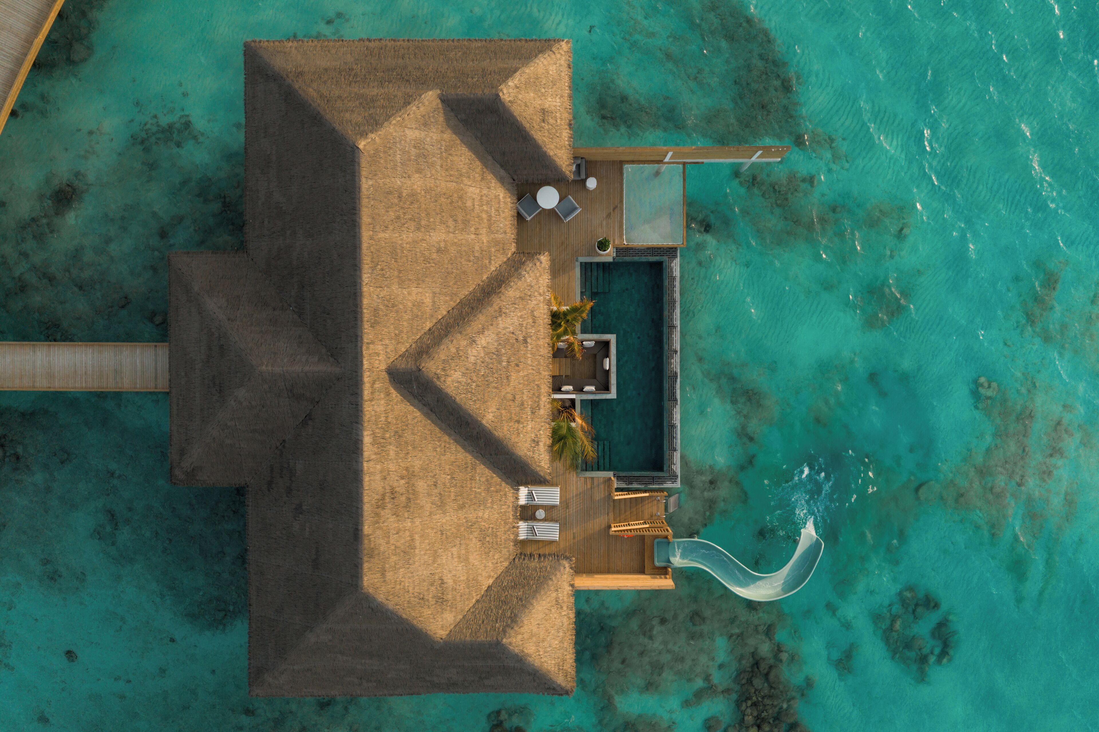 Vista aérea de un complejo turístico con piscina, sillones de descanso y una estructura de techo de paja.