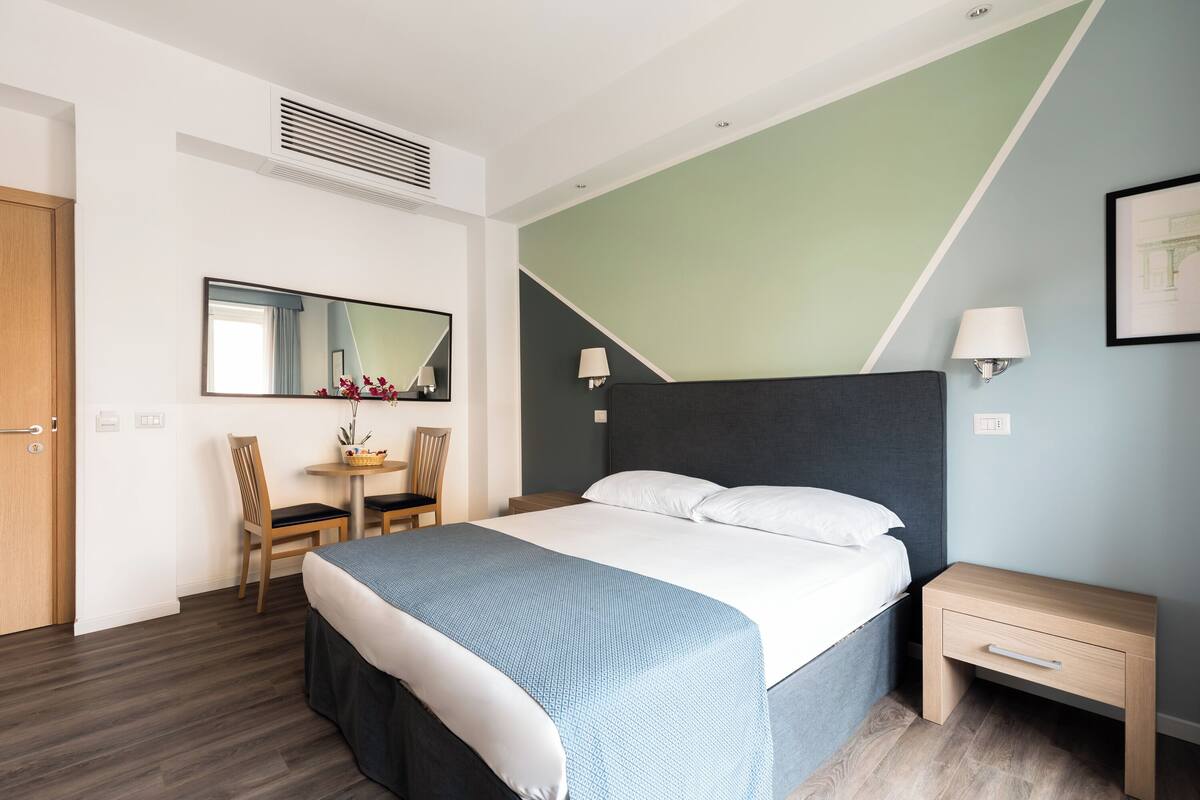 Una habitación de hotel moderna con cama, mesita de noche, silla, escritorio y un espejo grande.