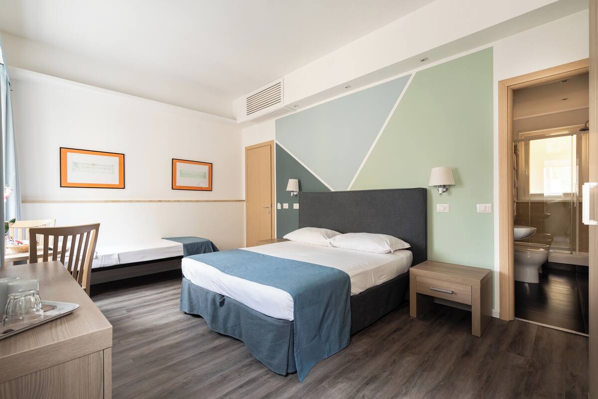 Habitación de hotel con cama, mesita de noche, silla, escritorio y vistas al baño.