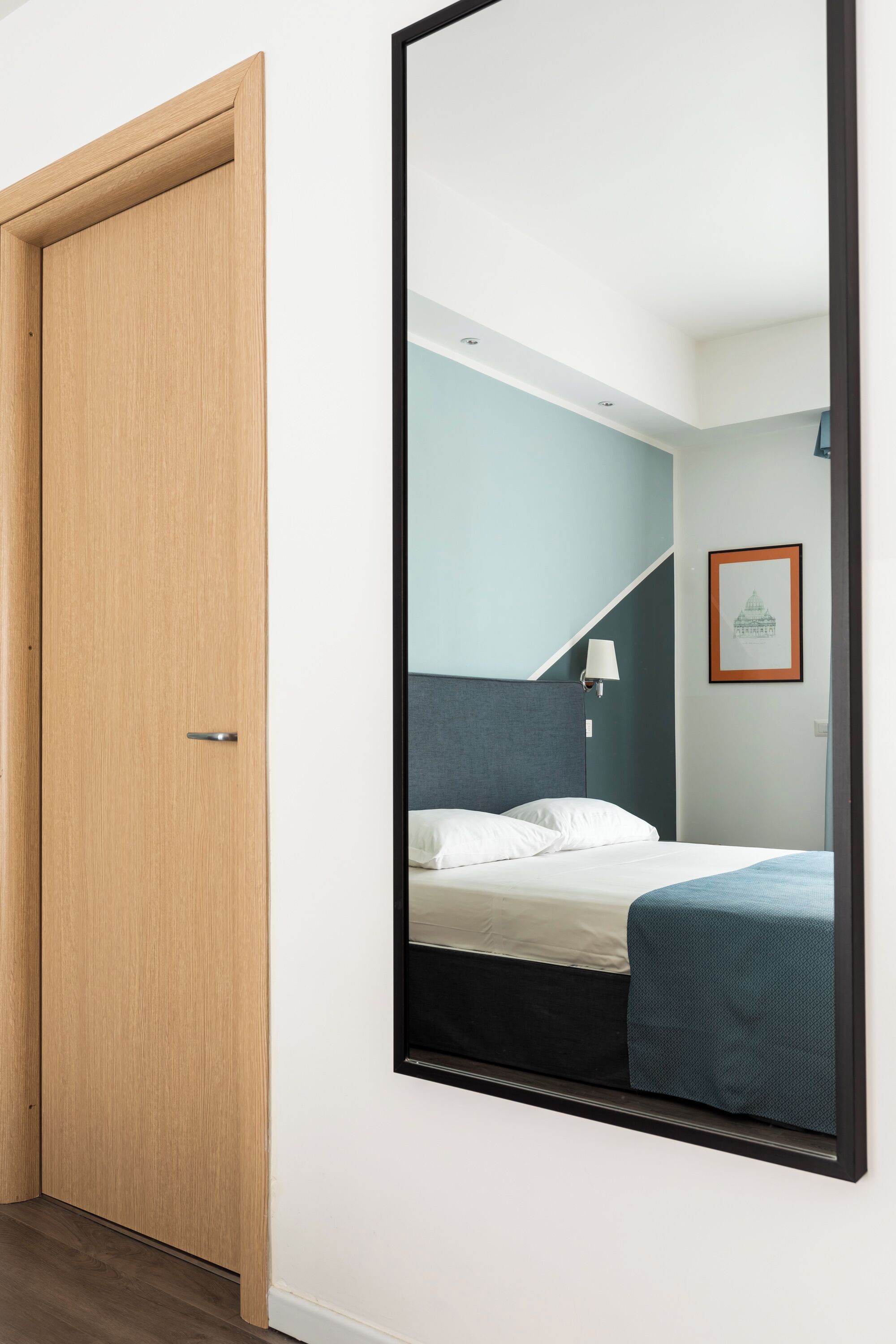 A hotel room with a wooden door, a bed with white and blue bedding, a wall-mounted mirror, and a framed picture on the wall.