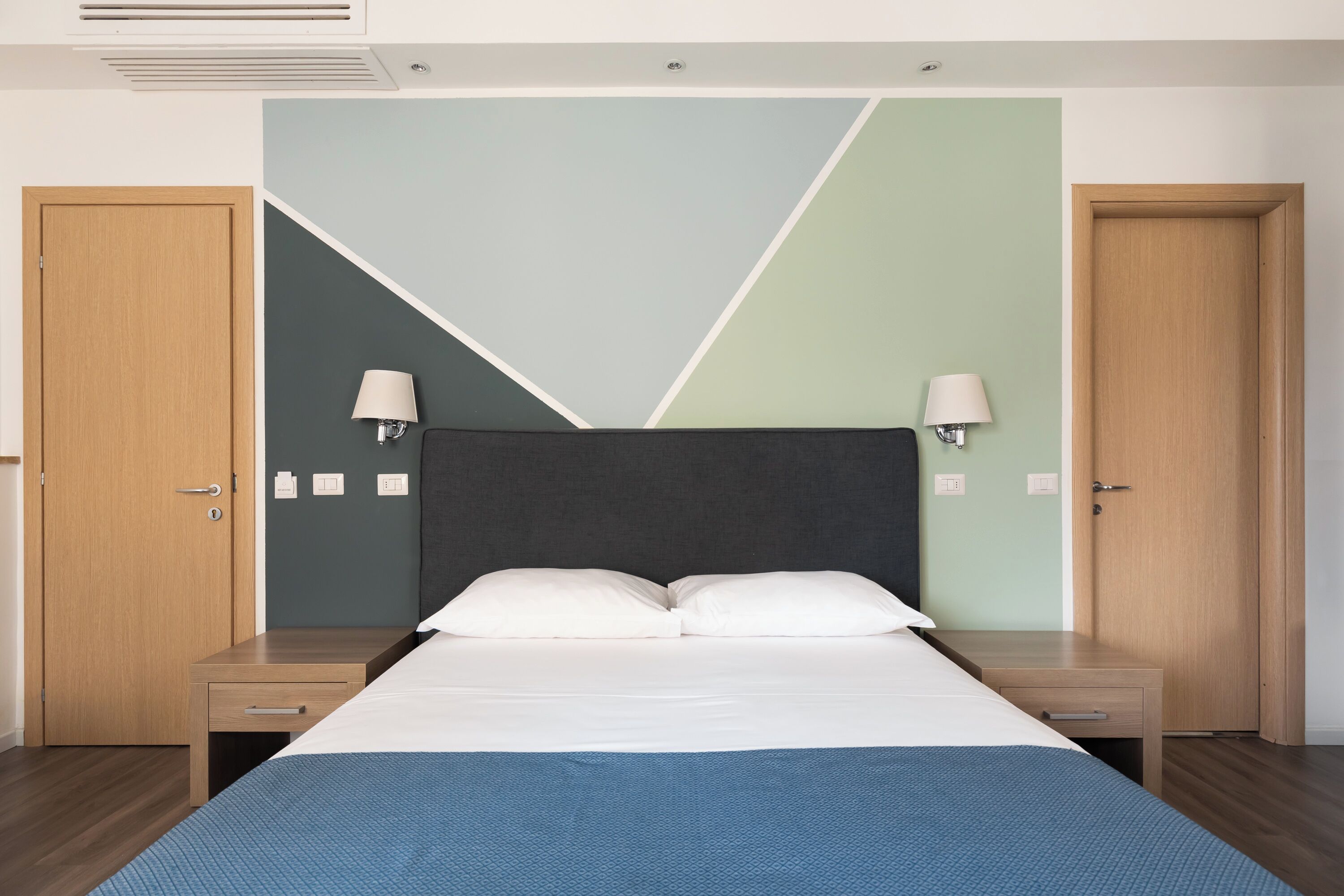Une chambre d’hôtel avec un lit, deux tables de chevet, une tête de lit et deux portes.