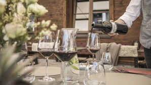 A person pouring wine into a glass at a table with several empty wine glasses and a vase of flowers. - Hotel Lenz (See)