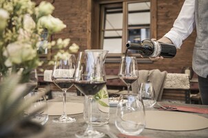 A person pouring wine into a glass at a table with several empty wine glasses and a vase of flowers.