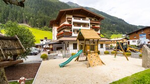 A playground with a slide and climbing structures, surrounded by a building with balconies and a mountain backdrop. - Hotel Lenz (See)