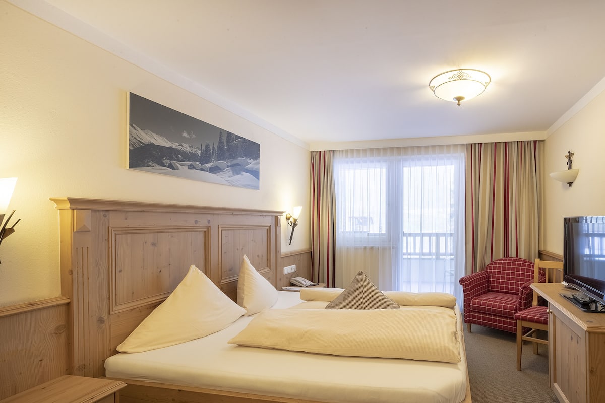 Habitación de hotel con cabecera de madera, cama con ropa blanca, televisor, escritorio con silla y ventana con cortinas.