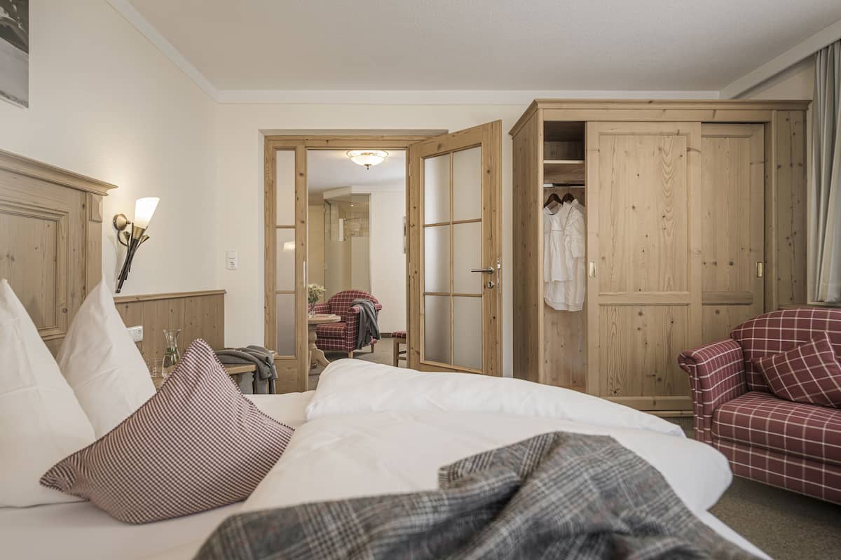 Una habitación de hotel con una cama, un sillón rojo a cuadros, un armario de madera y una mesita de noche con una lámpara.