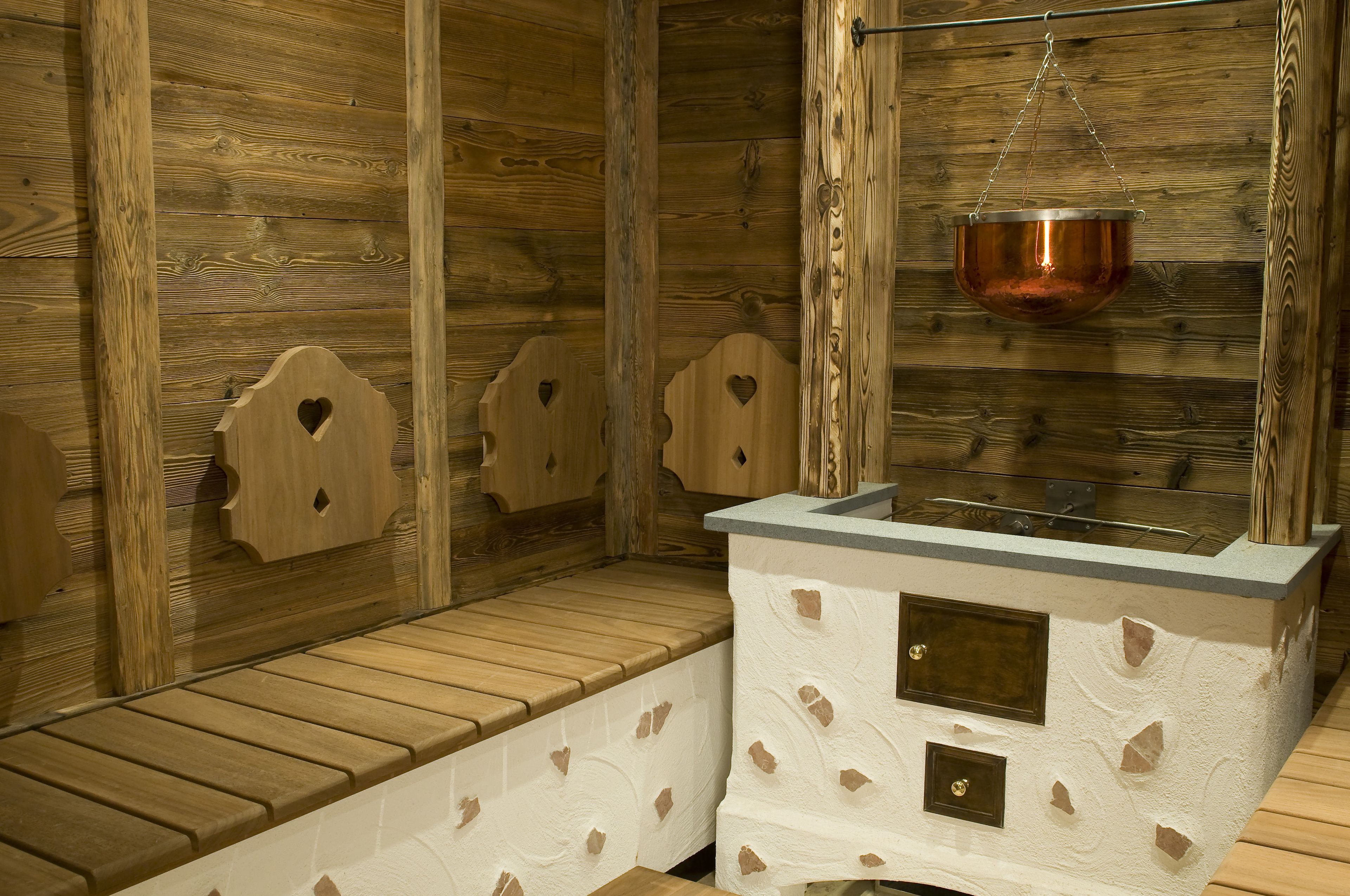 A sauna room with wooden benches, a hanging copper basin, and a stone wall.
