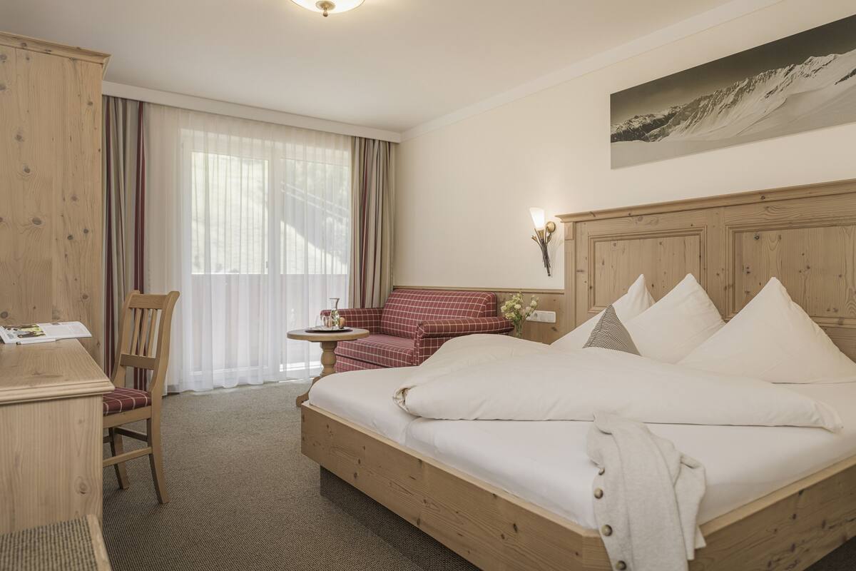 Habitación de hotel con cama de madera, un escritorio, una silla, una mesita y un ventanal con cortinas.