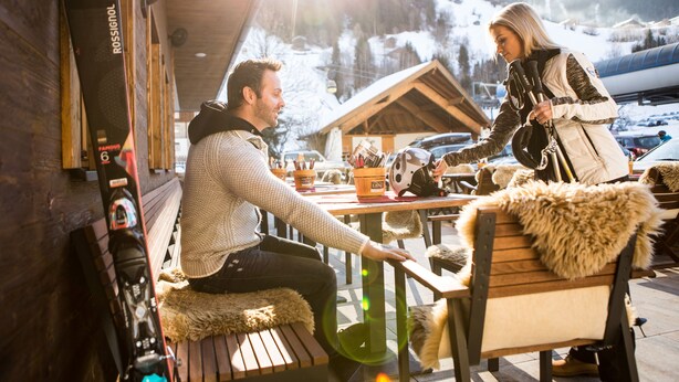A wooden bench with a fur throw, a ski rack, and a view of a snowy mountain in the background. - Hotel Lenz (See)