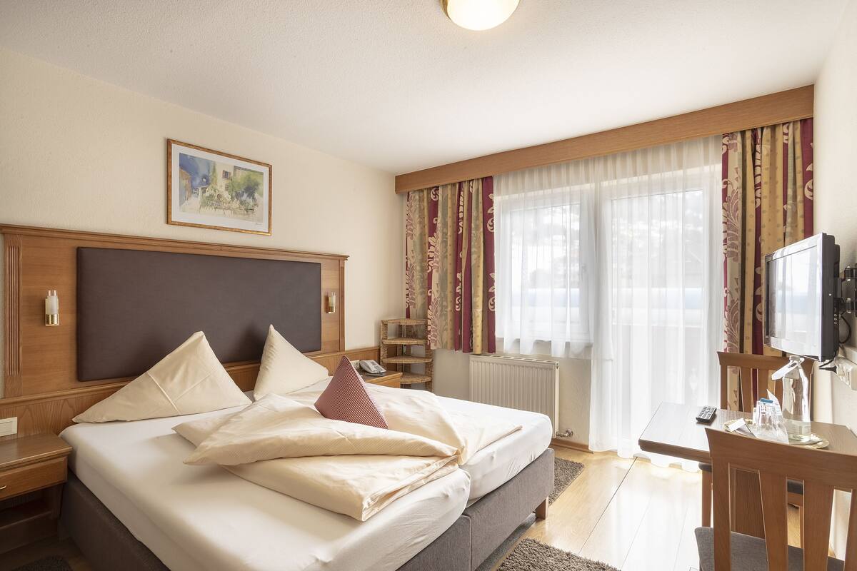 Una habitación de hotel con una cama grande, un cabecero de madera, una mesita de noche, un televisor y una ventana con cortinas.