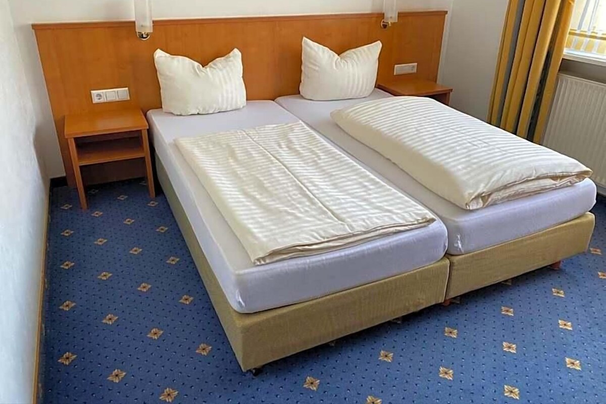 A hotel room with two beds, wooden headboards, bedside tables, and a blue carpet with a pattern.