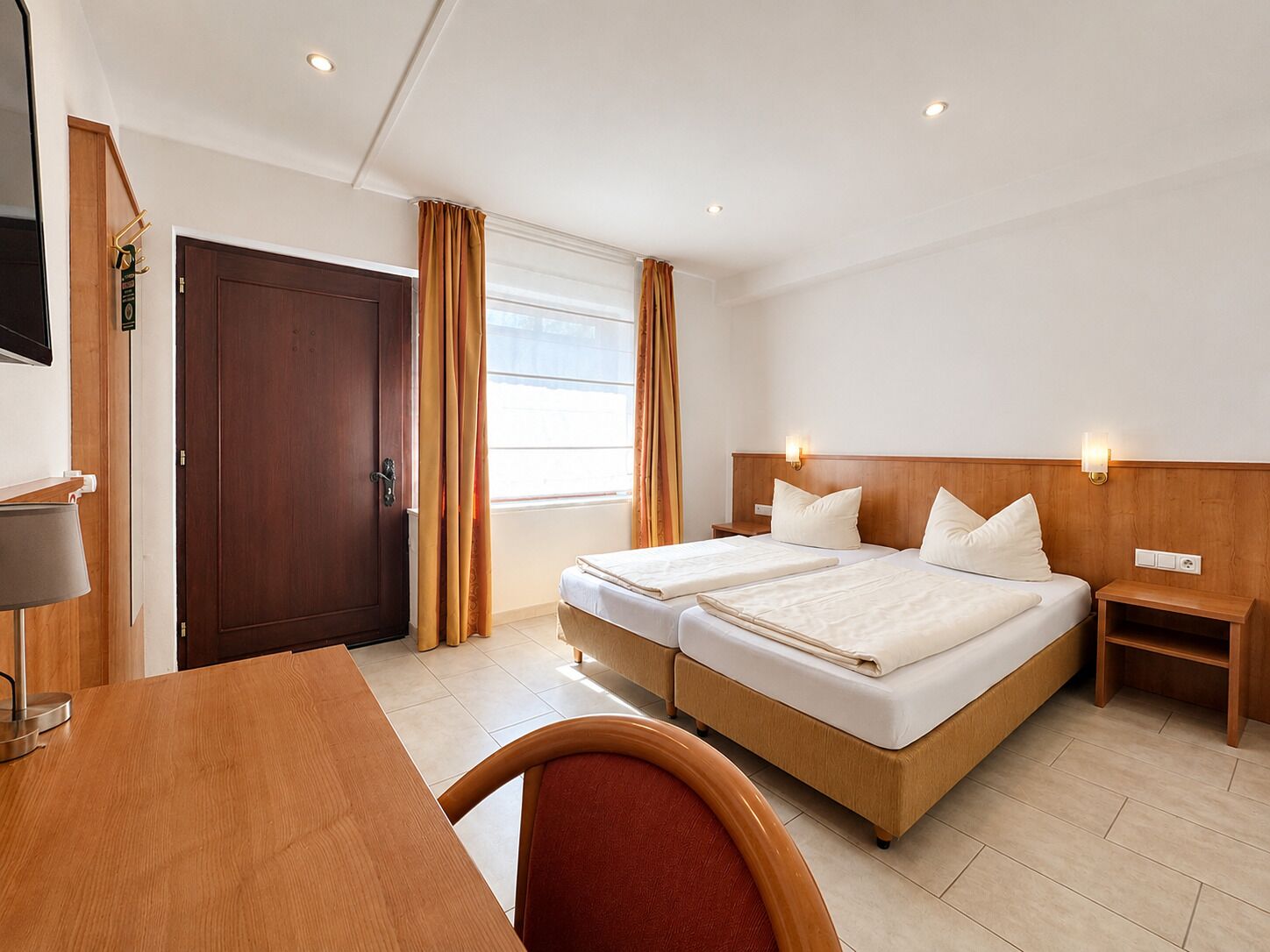 A hotel room with two beds, a desk, a chair, a TV, and a window with curtains.