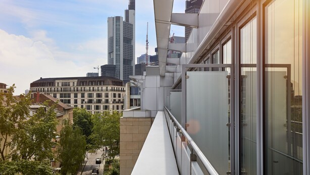 Un bâtiment moderne doté de grandes fenêtres et d’un balcon offrant une vue sur un paysage urbain avec de hauts immeubles et des grues de construction.