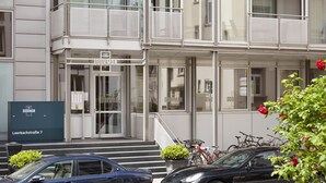 Un immeuble avec une pancarte indiquant « CONCIERGE » et « Leerbachstrasse 7 ». Deux voitures sont garées devant, et des vélos sont posés sur le trottoir.
