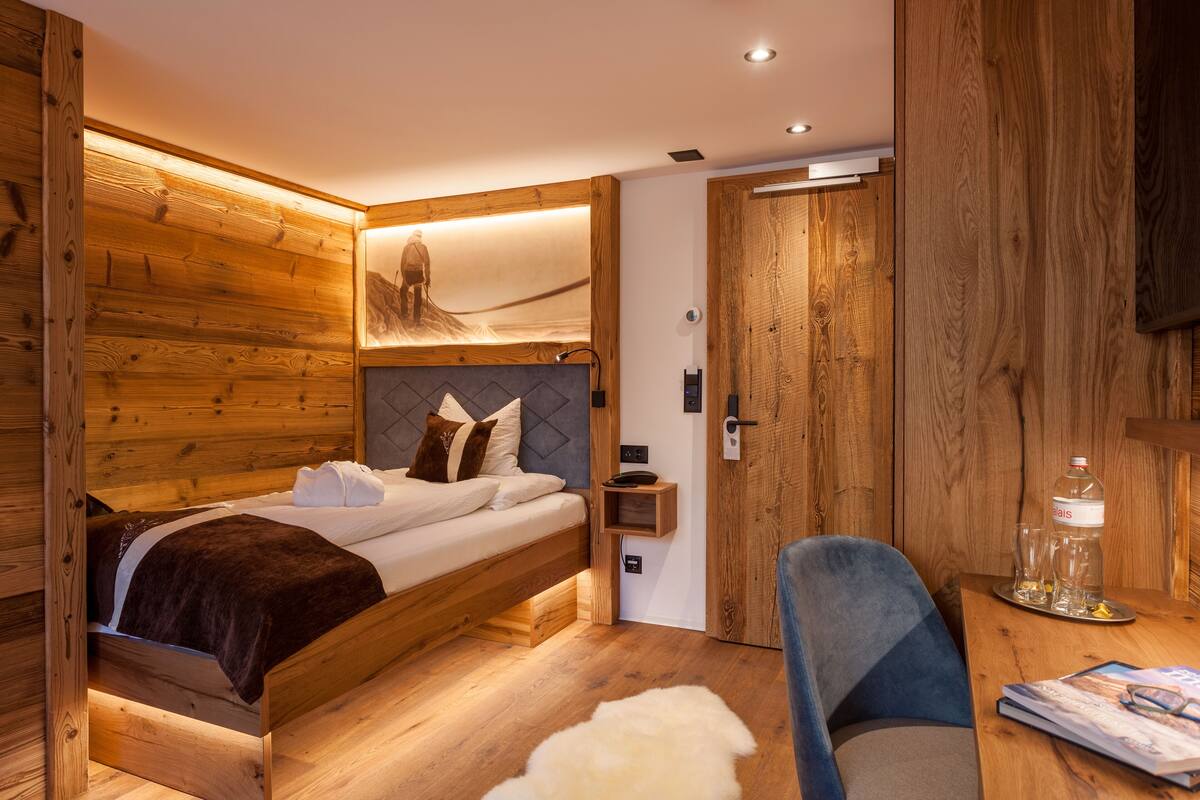 Ein holzmöbelgestaltetes Schlafzimmer mit einem Bett, einem Sessel, einem Tisch mit Gläsern und einer Flasche sowie einem an der Wand befestigten Regal.