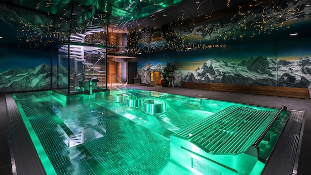 Un espace piscine intérieur moderne, doté d’une paroi vitrée, d’un coin salon et d’une fresque représentant des montagnes.