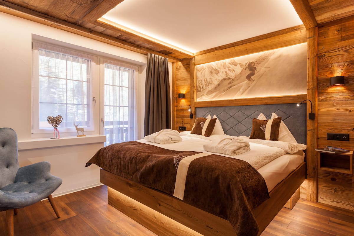 Ein schlafzimmer mit hölzernem Gestell, einem großen Bett, einem Fenster mit Vorhängen, einem Sessel und einer Wandleuchte.