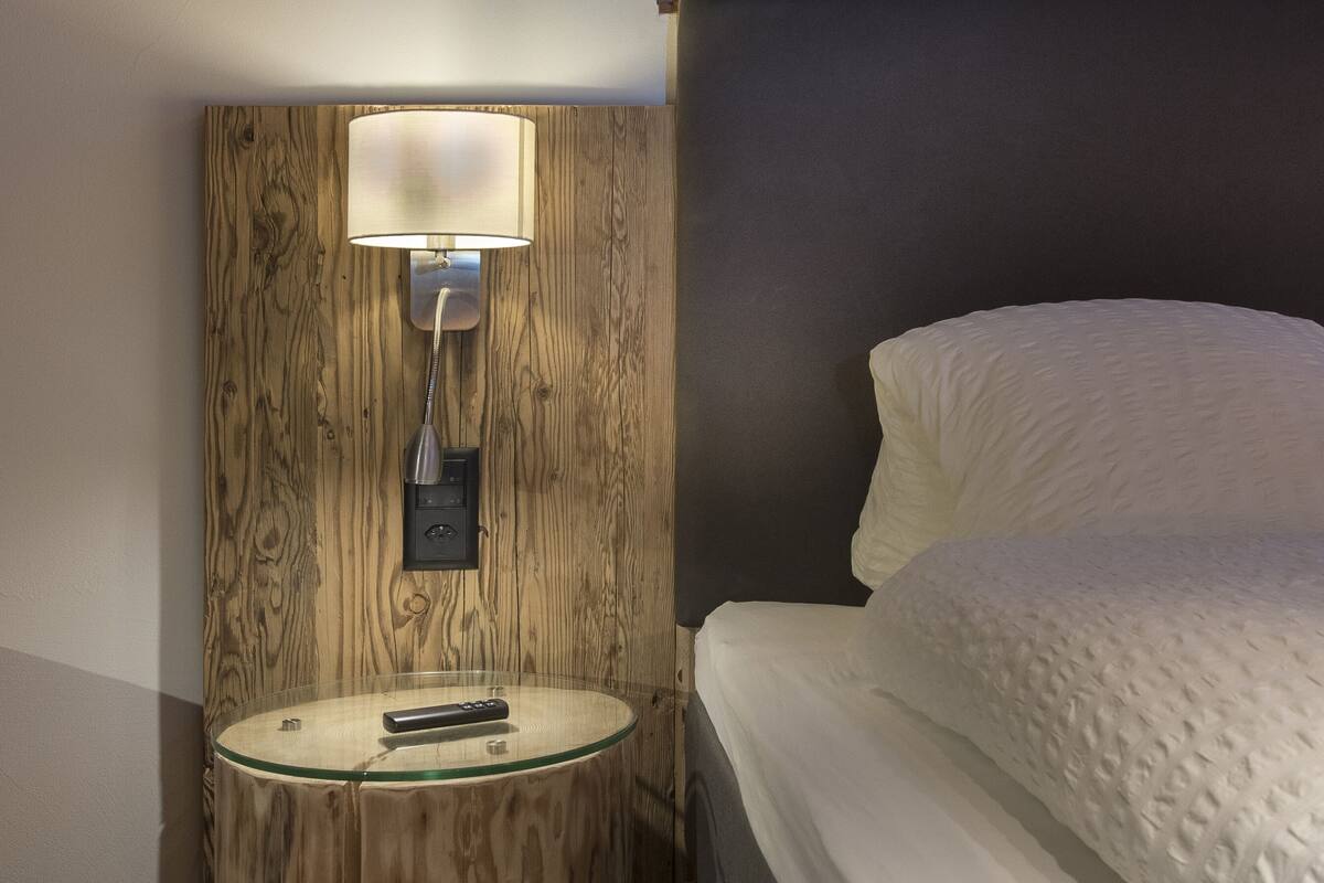 Ein ordentlich bezogenes Bett mit weißer Decke und Kissen, ein runder Nachttisch mit Lampe sowie ein kleiner Holzschemel.