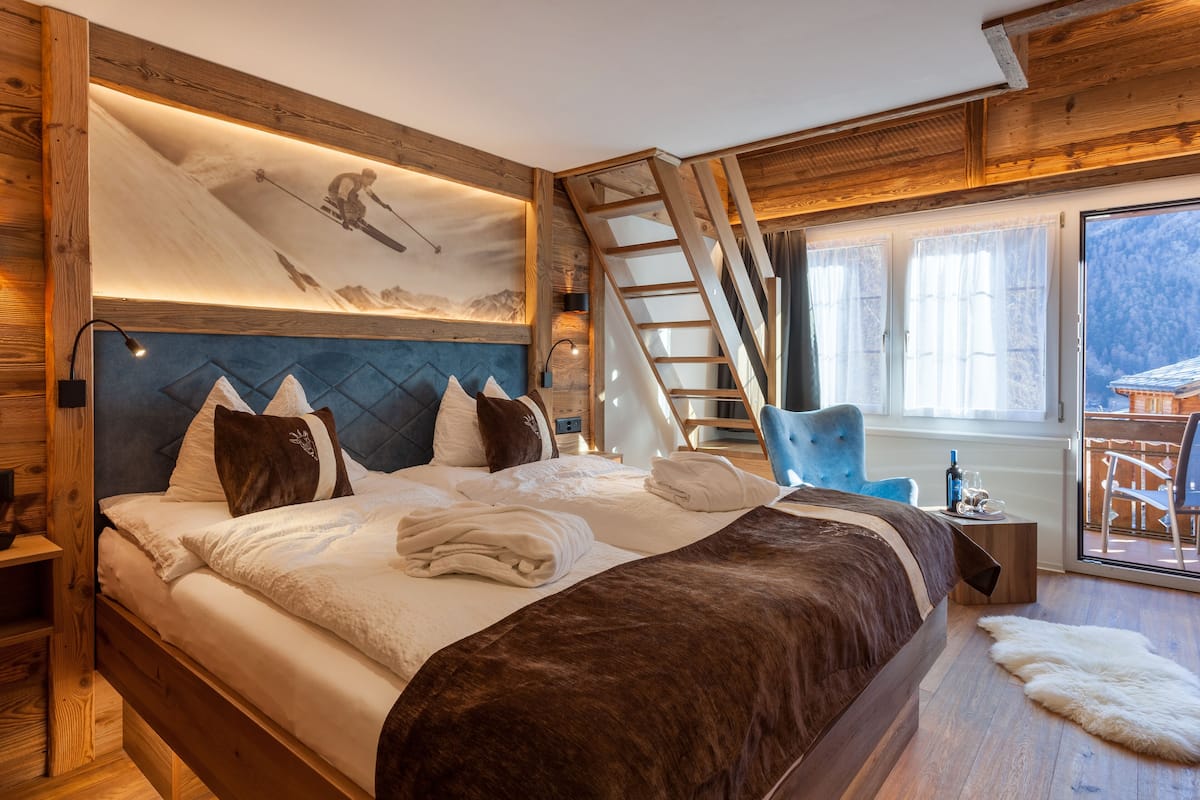 Ein Schlafzimmer mit einem großen Bett, einem Holzkopfende, einer Leiter, einem blauen Stuhl und Blick auf einen Balkon.