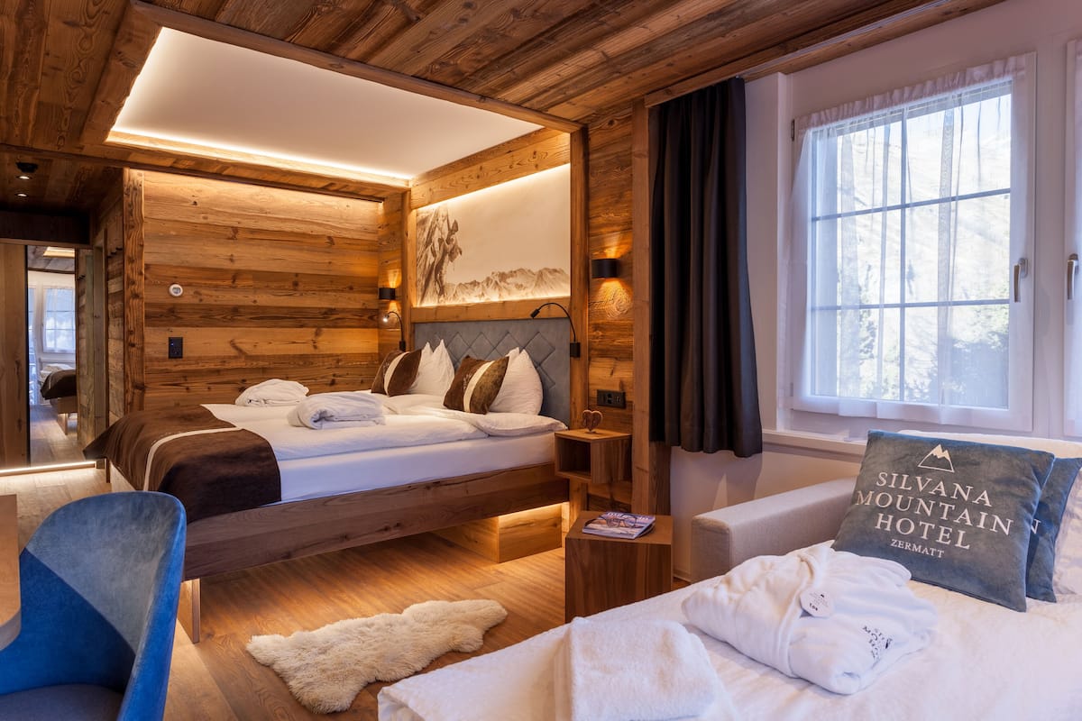 Ein schlicht eingerichtetes Schlafzimmer im Holzstil mit Bett, Sessel, Nachttisch und einem fenster mit Vorhängen.