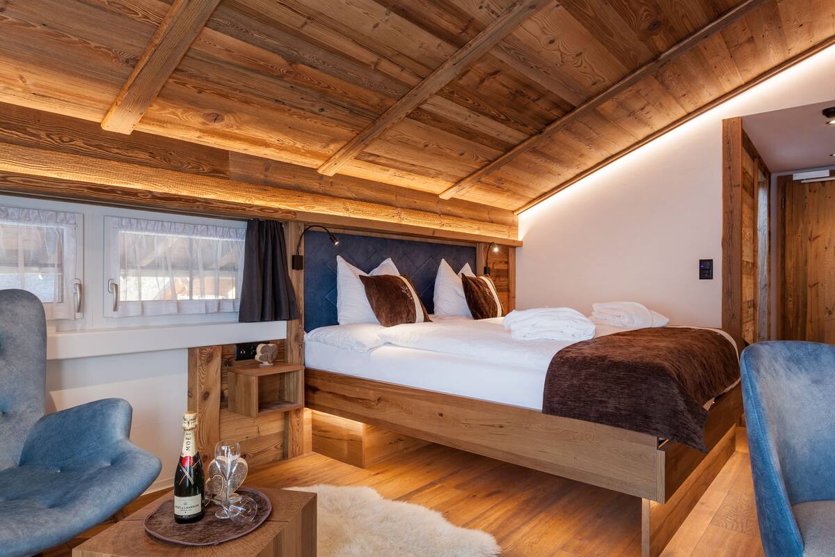 Ein schlafzimmer mit Holzrahmen, einem Bett, zwei Stühlen, einem kleinen Tisch mit einer Flasche und Gläsern sowie einem Fenster mit Vorhängen.