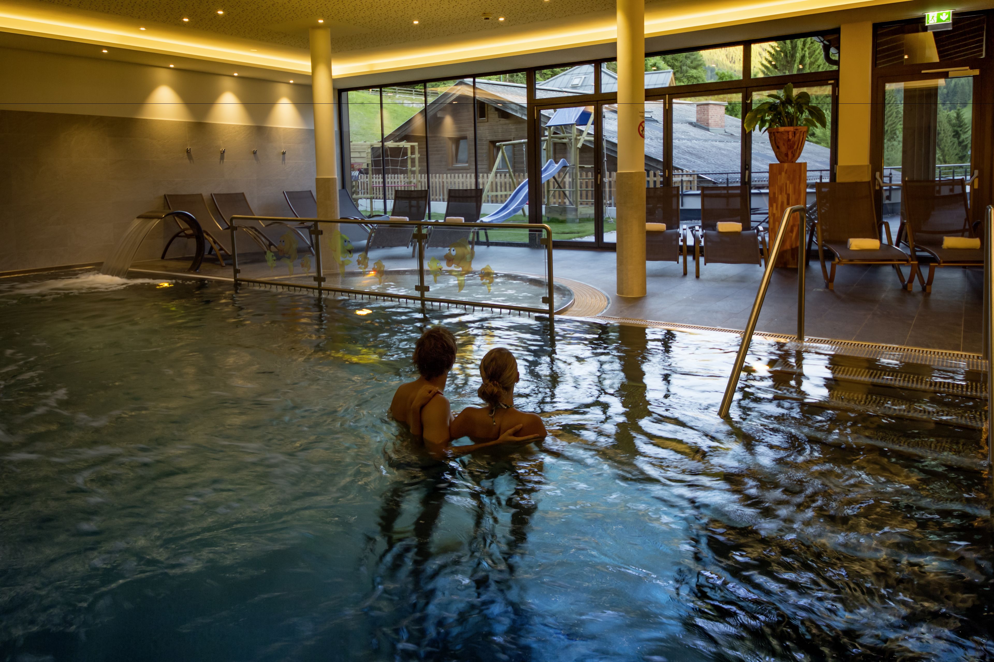 An indoor swimming pool with two people in the water, a slide, and a view of a wooden building outside.