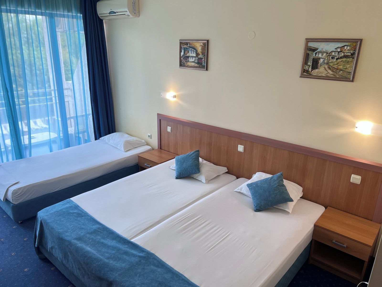 Habitación de hotel con dos camas, cabecero de madera, mesitas de noche, lámparas de pared y dos cuadros.