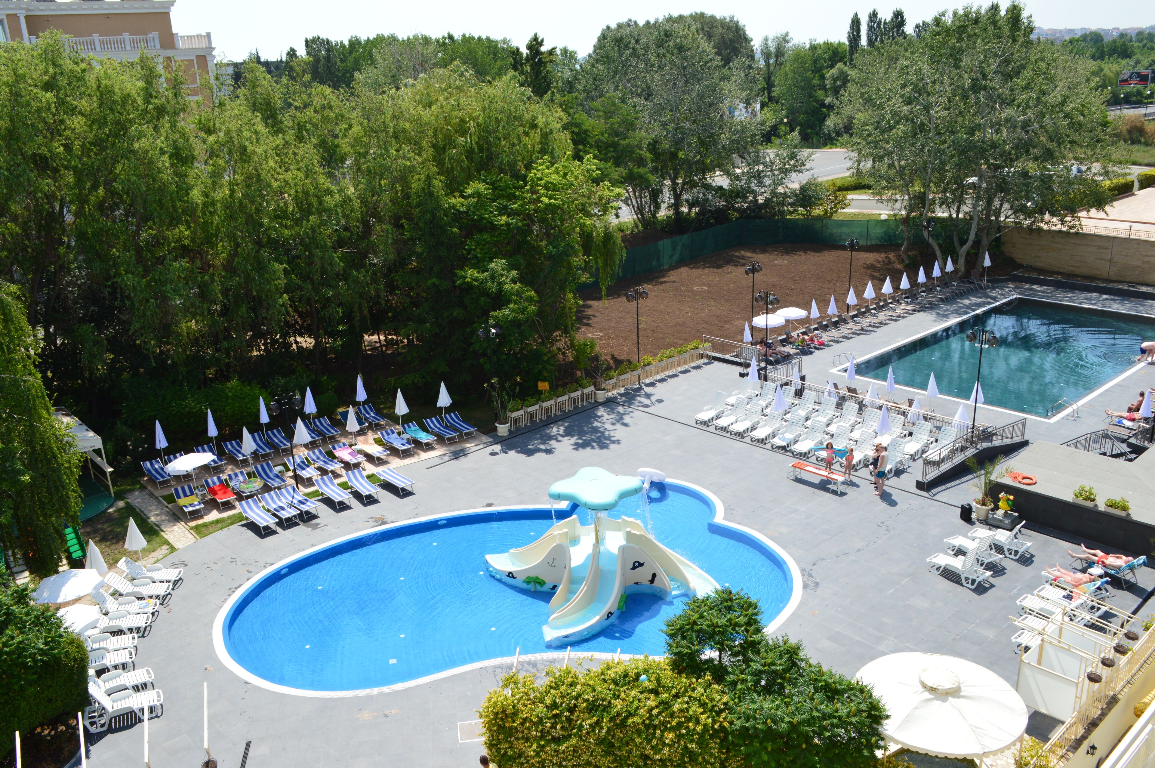 A swimming pool with a water slide, surrounded by lounge chairs and umbrellas, with trees and a building in the background.