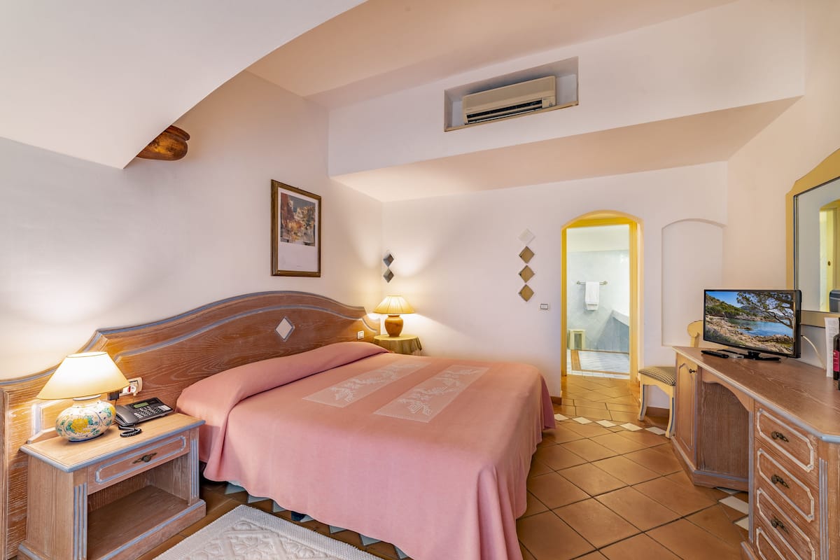 Un dormitorio con cama, lámparas de noche, televisión, cómoda y un baño visible a través de un arco.
