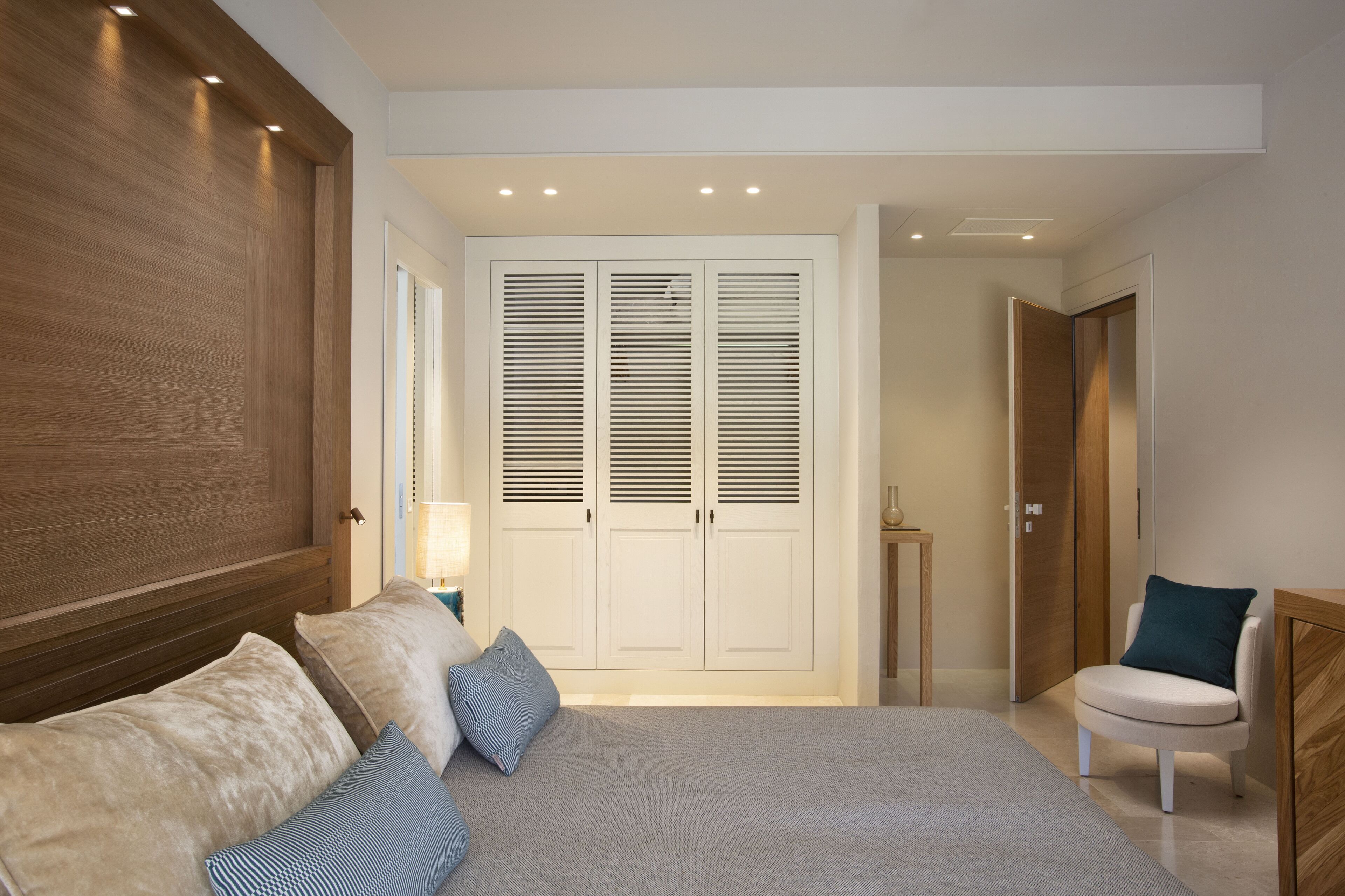 A bedroom with a large bed, a wooden headboard, a wardrobe with louvered doors, a bedside table with a lamp, and a chair with a cushion.