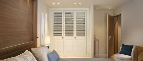 A bedroom with a large bed, a wooden headboard, a wardrobe with louvered doors, a bedside table with a lamp, and a chair with a cushion.