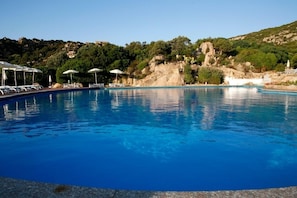 Une piscine d’un bleu éclatant, dotée de chaises longues et de parasols, entourée d’un paysage rocheux et de verdure.